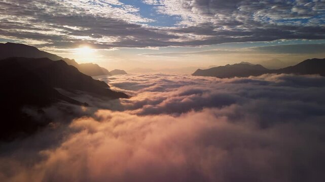 Aerial drone view of golden sunrise above a vast sea of clouds in the mountains. Soft morning light illuminates layered peaks rising through thick mist and drifting cloud inversion across the valley