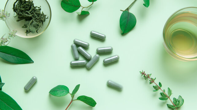 Flat lay of NMN supplement capsules with herbal tea and green leaves on a pastel green background. Wellness still life representing longevity, cellular energy, NAD+ metabolism and healthy aging