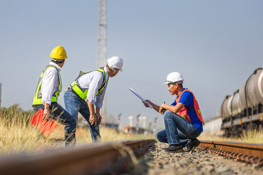 Professional engineering team inspecting railway track and infrastructure for safety and maintenance, Engineers reviewing site plans on railway track near freight train during field inspection
