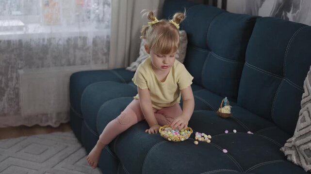 Soft light fills cozy living room. Toddler sits barefoot on plush sofa. Child wears yellow shirt, plays with sweets. Small hands reach for colorful candies scattered around