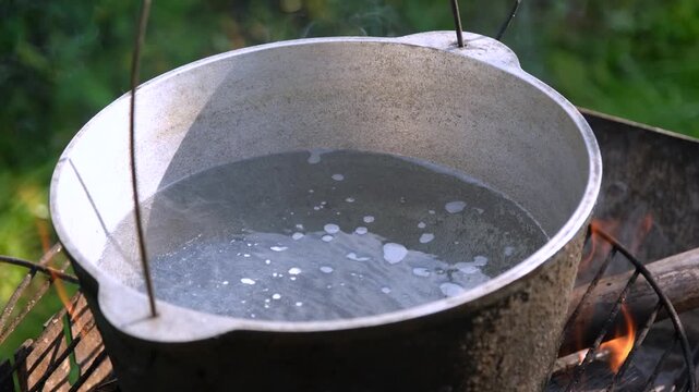 Close-up of water boiling with steam in a metal cauldron over an open campfire flame outdoors.