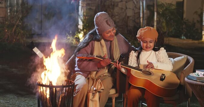 Indian elder woman lady sitting chair little kid girl hold guitar sing song enjoy night bonfire have fun joy happy relax old aged mom child learn music talk speak spend family time home garden place