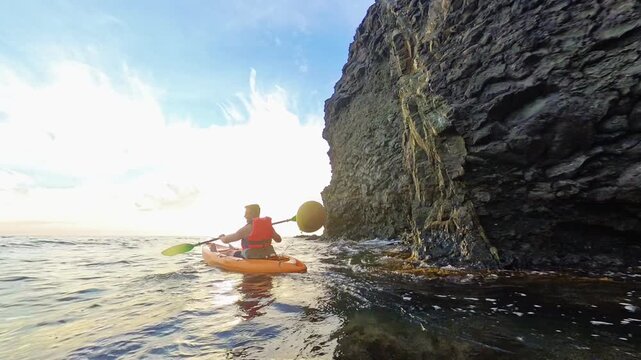 Kayak ocean adventure man paddling an orange kayak in the sea next to a giant rocky cliff during a golden sunset