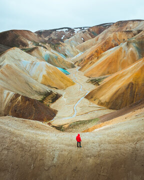 Aerial view of vivid, otherworldly mountainsides painted in hues of gold, cream, and turquoise, a tiny figure in red stands in awe, Rangarthing ytra, Iceland.