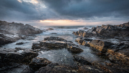 Fototapeta premium Dramatic Coastal Landscape with Crashing Waves and Rugged Rocks Under a Stormy, Overcast Sky at Sunset or Sunrise, Evoking a Powerful and Serene Mood