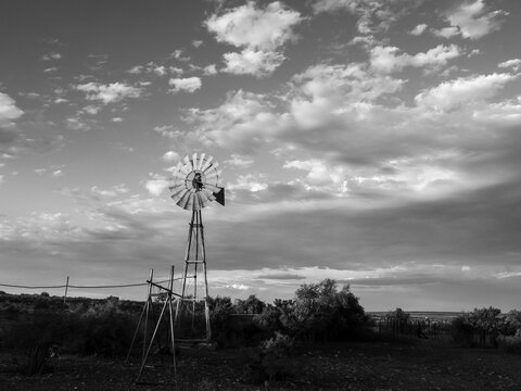 Black and white image of a windmill, blades spinning in the wind, pumping for water, in a rural Karoo field or farmland with clouds in the background