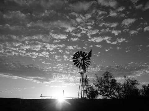Black and white image of a windmill, blades spinning in the wind, pumping for water, in a Karoo field or rural farmland with clouds in the background, and artistic glare from the sun setting