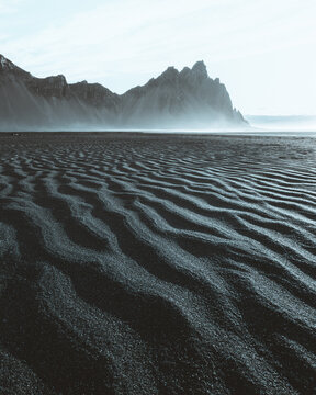 View of black sand beach with rhythmic patterns leading to distant, jagged mountains shrouded in mist, creating a stark, ethereal landscape, Sveitarfelagid Hornafjordur, Iceland.