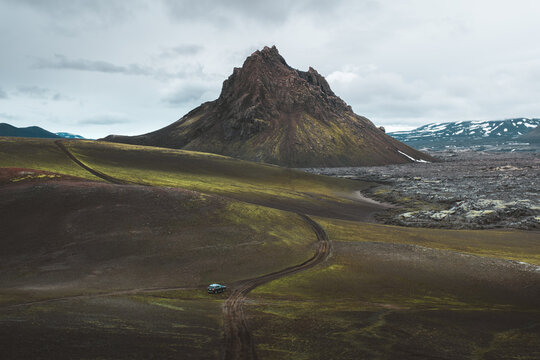 View of a lone car traversing a rugged path through a vivid landscape dominated by a sharp, rocky mountain under a cloudy sky, Rangarthing ytra, Iceland.