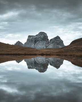 View of the majestic mountain mirrored in the serene lake under a cloudy sky, contrasting with the brown field, San Vito di Cadore, Veneto, Italy.
