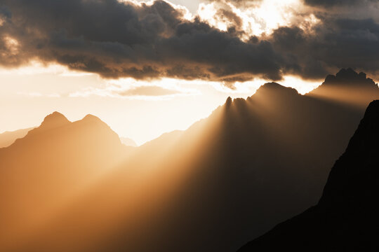 View of radiant sunbeams pierce through dark, brooding clouds, illuminating the majestic, silhouetted peaks of the Dolomites, San Vito di Cadore, Veneto, Italy.