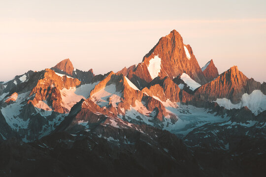 View of jagged, snow-capped peaks rise dramatically against a soft, orange-tinged sky, creating a stunning contrast of textures and tones, Obergoms VS, Switzerland.