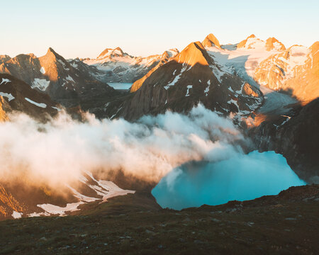 View of a serene turquoise lake nestled among jagged, snow-dusted peaks and swirling clouds bathed in the warm glow of sunrise, Obergoms VS, Switzerland.