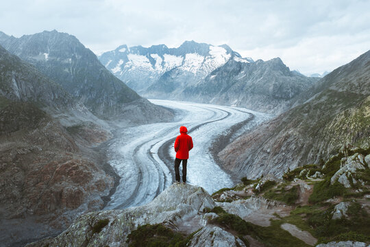 View of a lone figure in a vibrant red jacket standing atop a rocky precipice, overlooking the Aletsch Glacier snaking through the mountainous terrain, Torbel, Wallis, Switzerland.