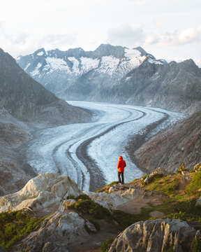 View of a solitary figure in a vibrant red jacket stands atop rugged terrain, gazing out over the Aletsch Glacier, Torbel, Wallis, Switzerland.