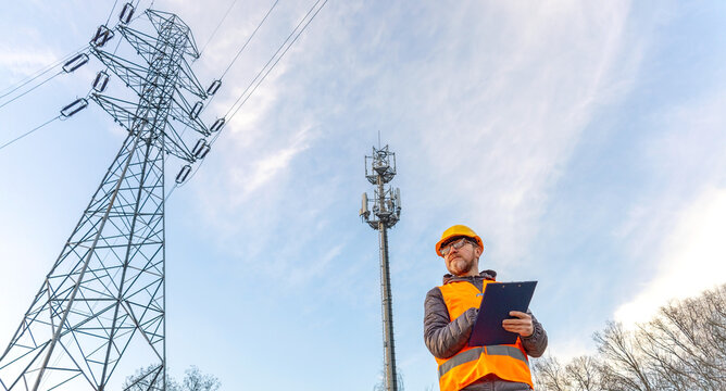 Electrical engineer inspecting power lines and a telecom tower. Professional inspector stands near high voltage transmission tower and a cellular 5g base station. Utilities infrastructure check.