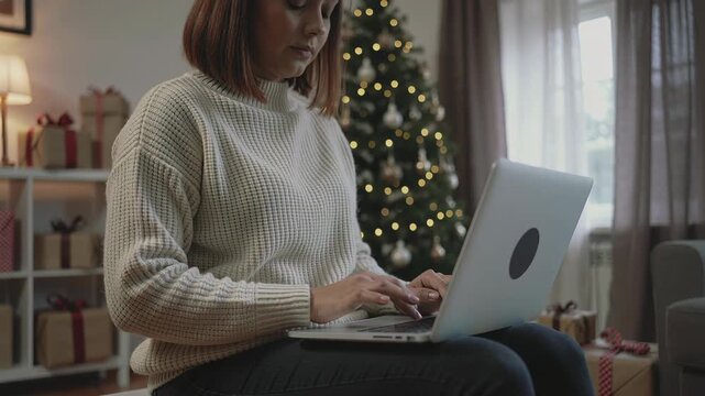 Woman typing on laptop near christmas tree in cozy living room setting