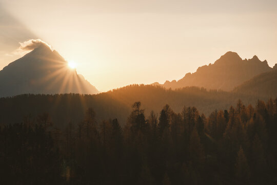 View of a golden sunrise bursting through a cloud-capped peak, casting long shadows across the forested valley and jagged mountain range, Cortina d'Ampezzo, Veneto, Italy.