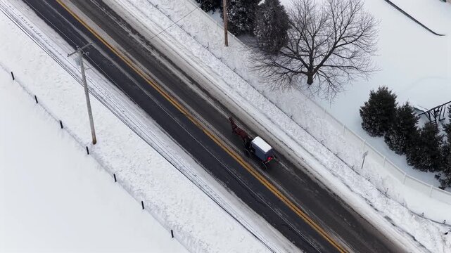 Aerial top down view of horse drawn carriage traveling along snowy rural road in Pennsylvania during winter, showing traditional Amish transportation crossing quiet countryside intersection.