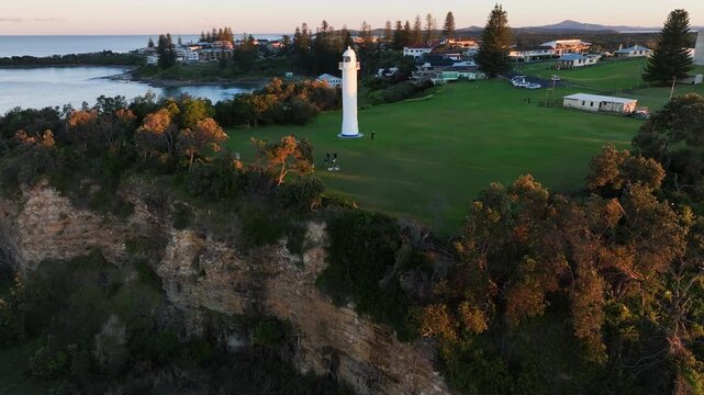 Drone glides toward Yamba Lighthouse at sunset, sweeping over vivid green grass and trees. Golden light washes the headland as the bay opens in the distance, calm water reflecting the fading sky.