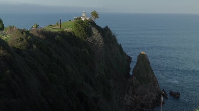 Stunning aerial footage of white lighthouse perched on rugged coastal cliff overlooking Atlantic Ocean in Candas, Asturias, Spain. Dramatic landscape with rocky formations and serene blue sea.
