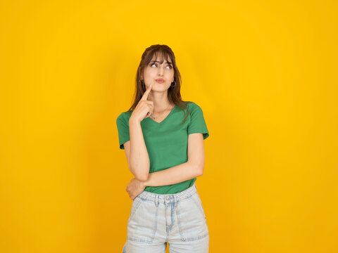 Thoughtful young woman touching her chin while looking aside, wearing green shirt and denim shorts, standing against bright yellow studio background with copy space. Thinking and decision concept.