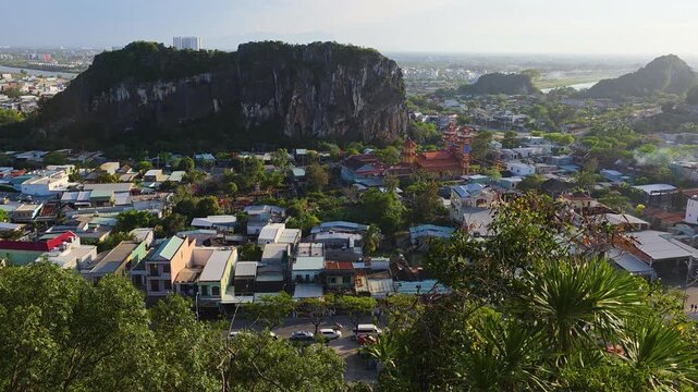 Elevated shot from Marble Mountains reveals dramatic sheer limestone cliff of Hon Moc Son towering above dense Da Nang residential rooftops with ornate Vietnamese temple gate, river and city skyline a