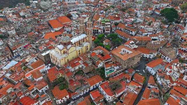 Tilt-up aerial view of the Santa Prisca temple revealing the city of Taxco, Guerrero, Mexico, with white colonial houses and tiled roofs on a sunny day.