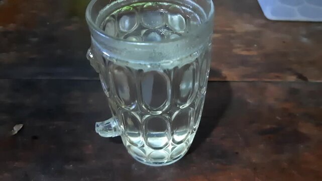 Glass mug containing a small amount of clear liquid sits motionless on a dark wooden surface as implied by the video footage.