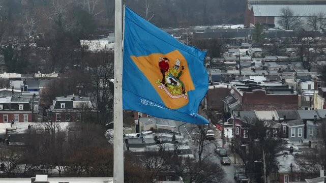 Aerial drone view of Delaware state flag waving in wind above snowy American city during winter. Historic rooftops and residential buildings appear in background under overcast sky. Close up shot: