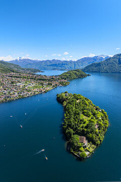 Aerial view of a lush island nestled in the blue waters of Lake Como, with Domaso town sprawling along the opposite shore, framed by distant snow-capped mountains, Domaso, Lombardy, Italy.