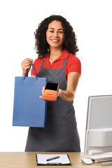 Female African-American cashier with payment terminal and shopping bag at checkout counter on white...