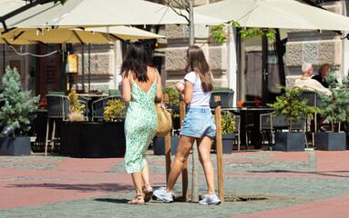 Back view of two attractive young women walking through a sunny city square past a café terrace with umbrellas and green plants © Popescu