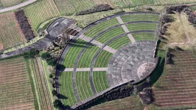 Aerial view of a semi-circular war memorial with rows of graves surrounded by fields and sparse trees creating a somber landscape, Mittelwihr, Grand Est, France.