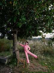 Naklejka premium Balance, outdoor, woman, playful, pink jumpsuit, tree, nature, posing in a countryside setting with sunlit meadow, capturing leisure and carefree athletic vibe in a scenic outdoor moment for stock use