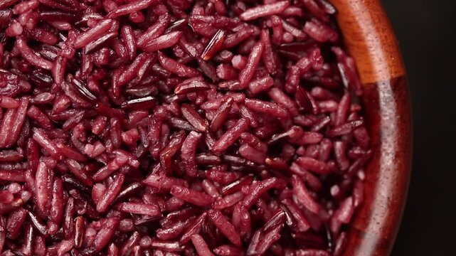 Cooked riceberry rice in a wooden bowl rotating slowly on black background. Top view shot