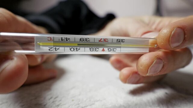 Male hands hold glass thermometer showing 39 degrees Celsius before one hand falls weakly. Close-up shot captures the details of the measurement and subsequent fatigue.