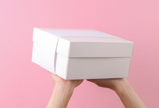 Female hands holding white premium cardboard box with paper stripe on pink background. Mockup for design.