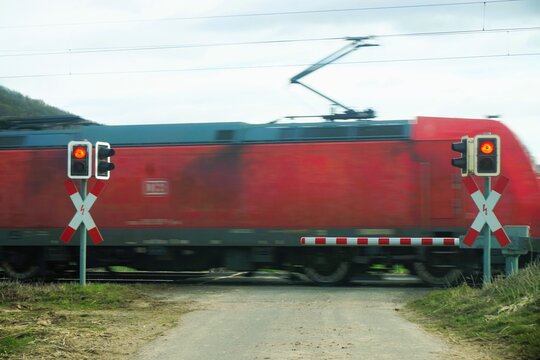 Bahn&uuml;bergang mit roten Ampeln und Schranke vor durchfahrendem roten Zug auf dem Land im Fr&uuml;hling