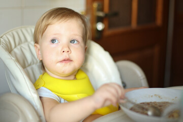 Fototapeta premium The little baby eats independently while sitting at the feeding table