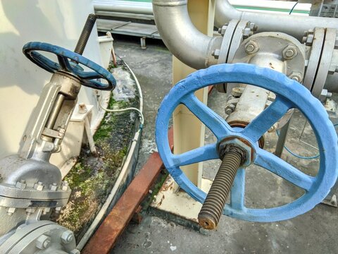 Industrial manual gate valves with blue handwheels on steel pipelines. Close-up of mechanical flow control systems at a factory storage tank area. Engineering infrastructure for fluid management.