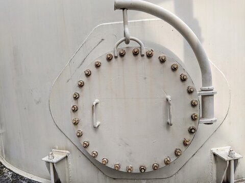 Industrial steel manhole cover on a storage tank with heavy-duty bolts, davit arm hinge, and handles. Industrial pressure vessel access hatch for maintenance and inspection.