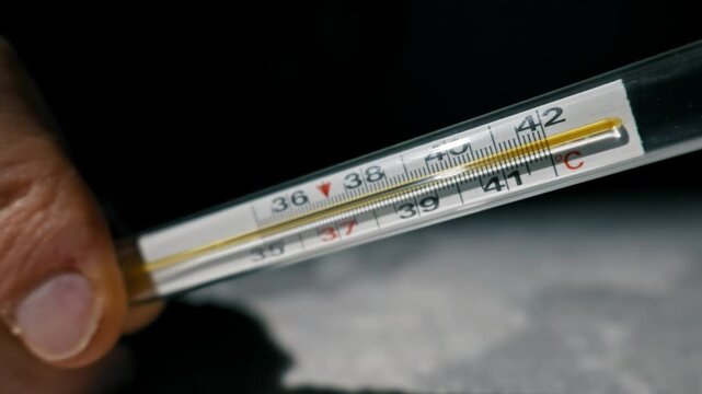 Male hand holds a classic glass mercury thermometer showing a dangerously high fever of 42 degrees Celsius. Extreme close-up shot focuses on the temperature scale against a dark background.