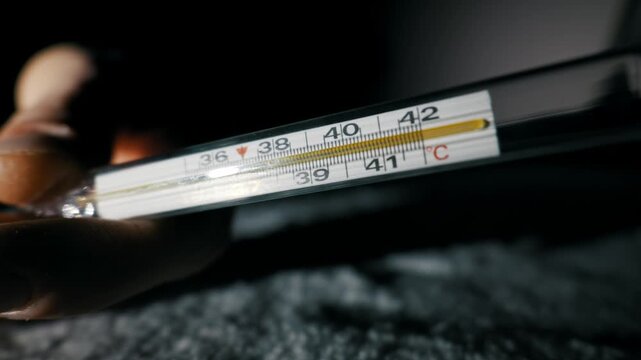 Person hand holds a glass mercury thermometer indicating a dangerous fever over 41 Celsius. Dramatic low-key lighting creates a serious and concerning mood in this close-up shot.