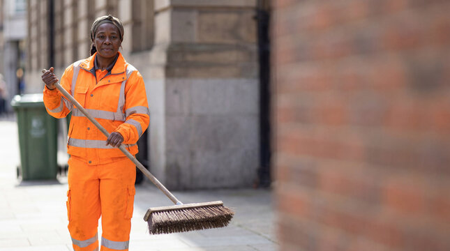 Female sanitation worker in orange high-visibility uniform holding a push broom. Black woman street sweeper working on a city sidewalk. Public service and manual labor concept