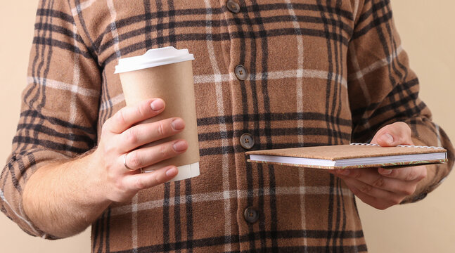 Man in flannel checkered shirt is holding eco friendly coffee cup and notebook on beige background