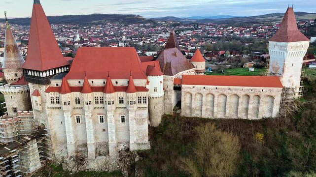 Aerial drone view of Corvin Castle in Hunedoara, Transylvania. Medieval Gothic fortress with red tiled roofs, stone towers, and dramatic defensive walls rising above the town