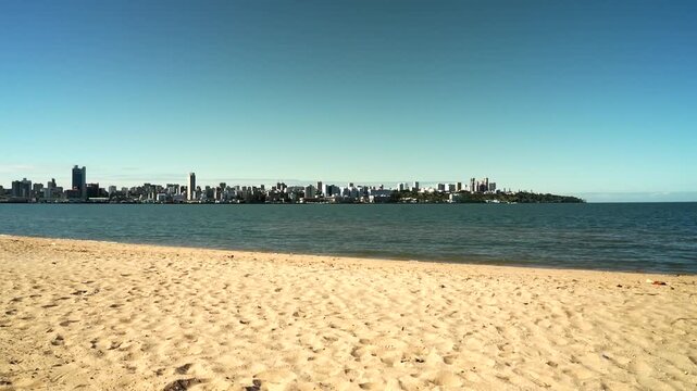 Wide-angle skyline of Maputo, Mozambique across Maputo Bay from sandy beach, cluster of high-rise towers and buildings on clear sunny day with blue sky and ocean.