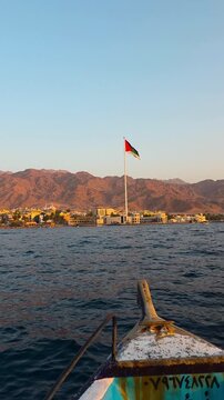 POV vertical footage from a glass boat approaching the Arab Revolt Flag on the Aqaba waterfront, Jordan, with calm sea, coastal cityscape and warm sunset light