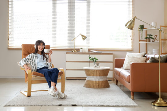 Young woman with headphones and tea cup sitting in leather armchair at home